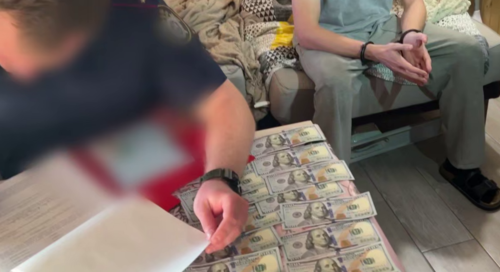 Силовик пишет протокол на разложенных долларах.Стоп-кадр видео Следкома России по Краснодарскому краю от 25.09.25, https://t.me/kubansledcom/6066
